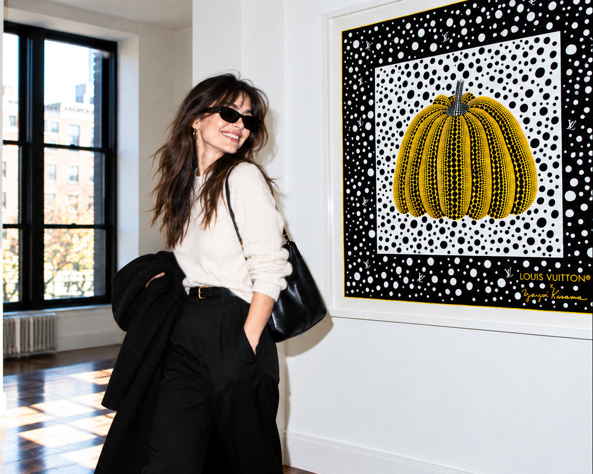 A woman wearing black sunglasses, a cream long-sleeve top, and black wide-leg trousers stands in a sunlit gallery room next to a framed art print featuring a yellow cactus on a black and white spotted background.