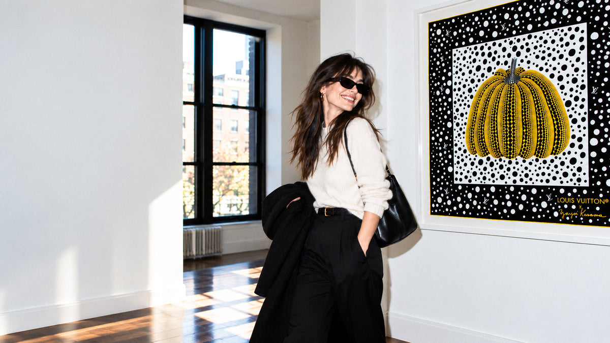 A woman wearing black sunglasses, a cream long-sleeve top, and black wide-leg trousers stands in a sunlit gallery room next to a framed silk scarf featuring a Louis Vuitton x Yayoi Kusama 90 Infinity Dots Pumpkin Scarf in black and yellow.