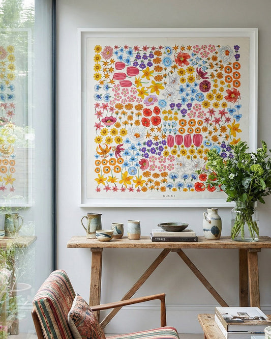 Colourful Gucci floral framed silk scarf artwork on a wall above a wooden table with decorative items. The artwork is hung in an elegant Sussex home on a wall next to a window to the garden.