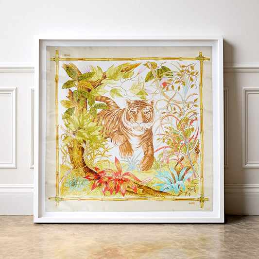 A framed art silk scarf featuring a detailed illustration of a tiger in a lush tropical  jungle, set in a white frame and leaning against a white panelled wall. Il Gattino translates from Italian as The Kitty or the Kitten. This beautiful Gucci print has been selected by Gucci's new Creative Director for 2026 Denma. The image itself is beautifully framed by a bamboo edging. 