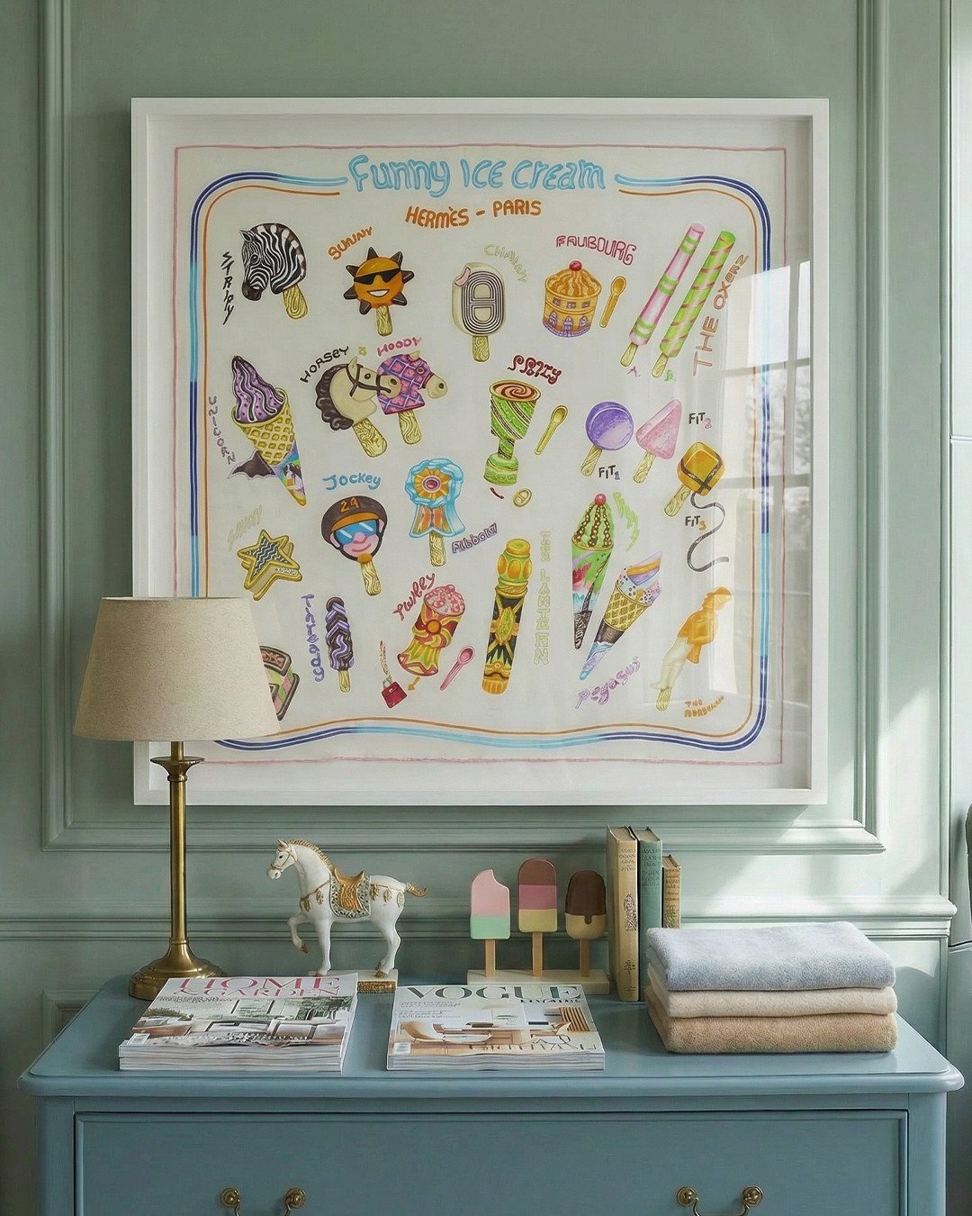 Framed children's ice cream artwork as a framed silk scarf, on a wall above a blue dresser with Vogue magazine, Home & Garden magazine, toy horse, ice cream toys, a lamp, and soft baby blankets.