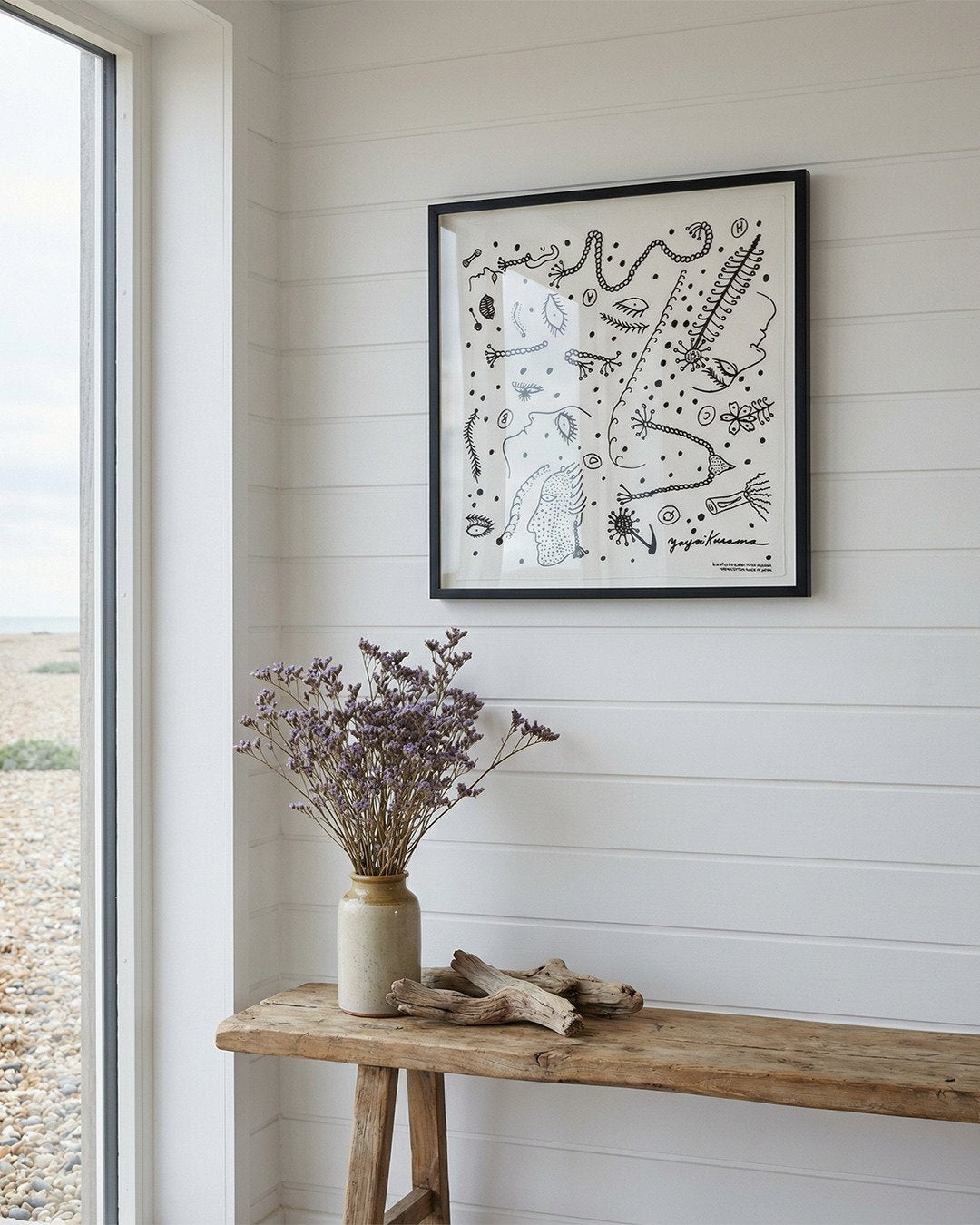 Framed Yayoi Kusama artwork of the Lammfromm Tokyo Black and White cotton textile scarf. The artwork is on a wall in a beautiful Sussex beachside home, above a wooden bench with a vase and dried flowers.