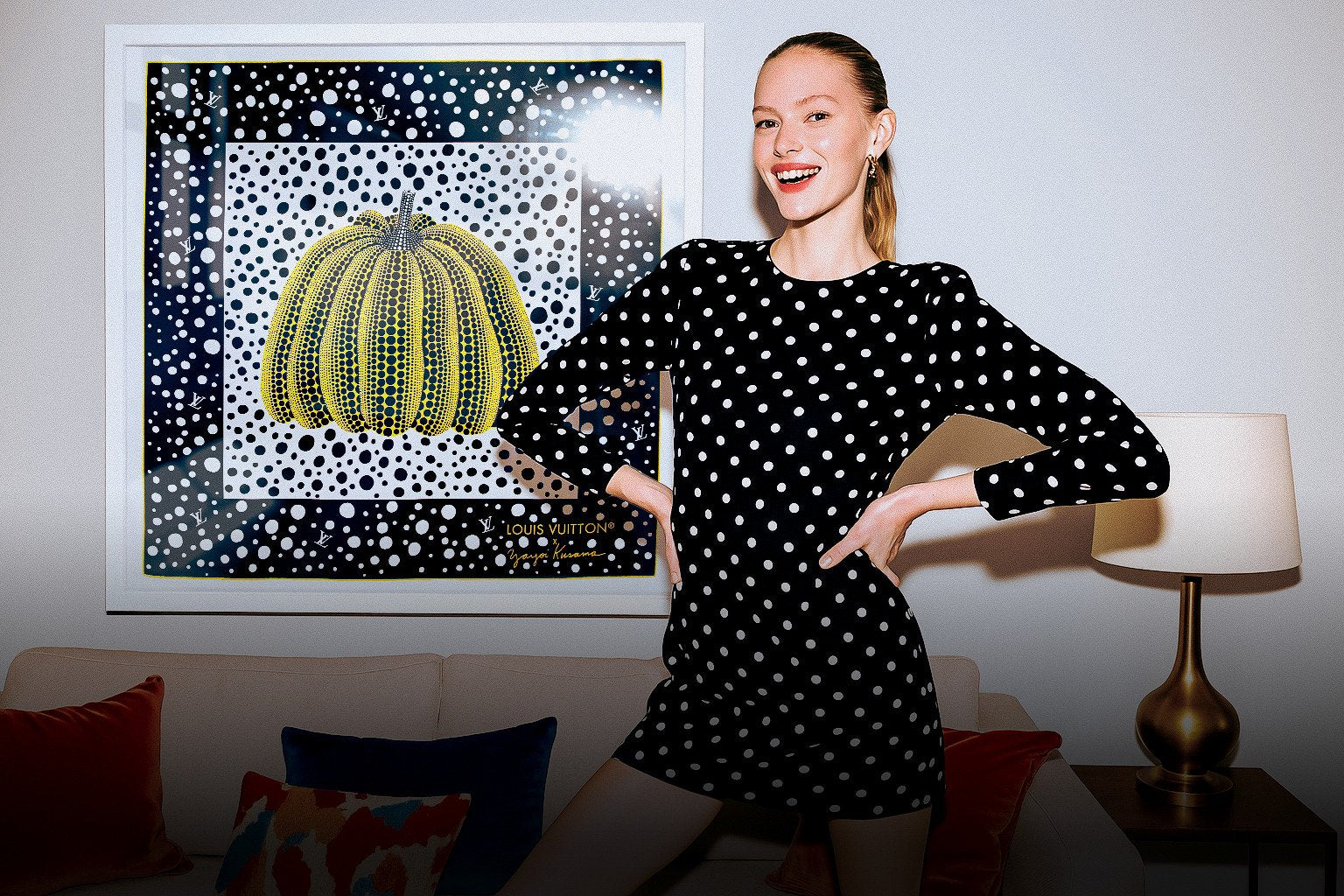 Model in polka dot dress posing in front of a framed Louis Vuitton x Yayoi Kusama yellow pumpkin silk scarf, styled in a modern interior with velvet cushions and brass lamp.